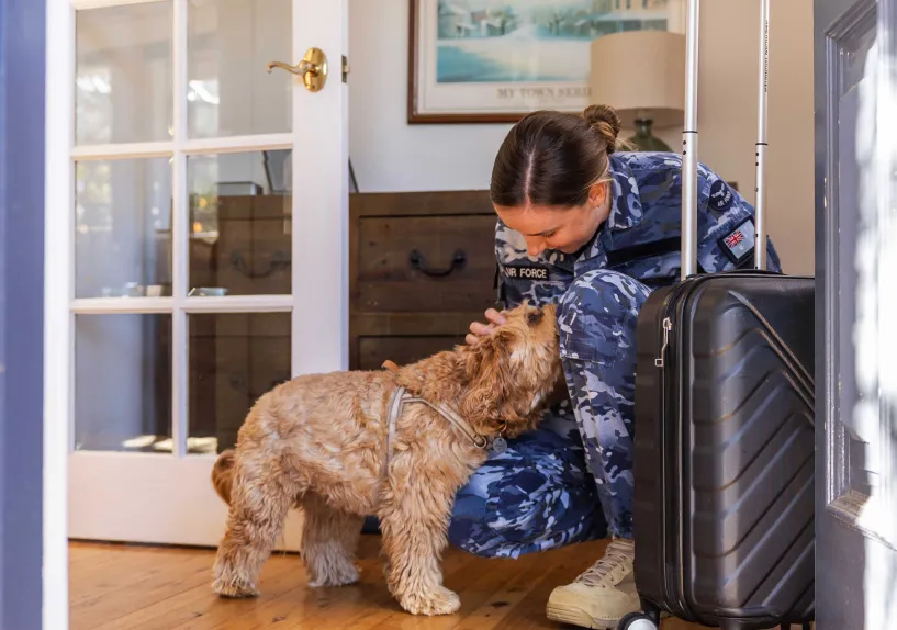 A person in an Australian Defence Force uniform kneeling to greet a dog beside a suitcase at home.