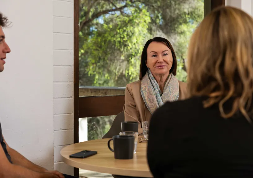 Three adults sitting around a table having a conversation over coffee in a casual indoor setting.
