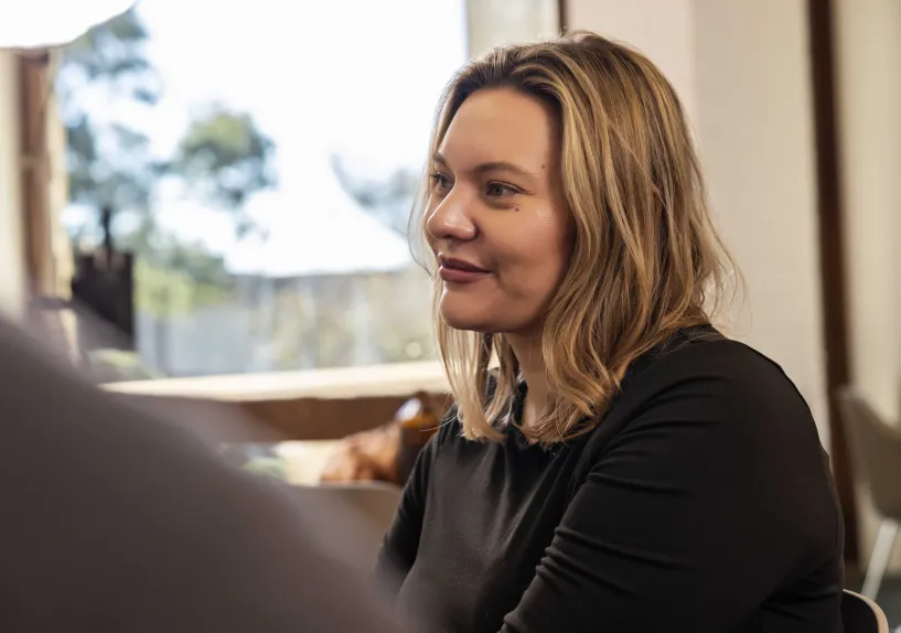 A person sitting indoors, facing and listening to someone in conversation.
