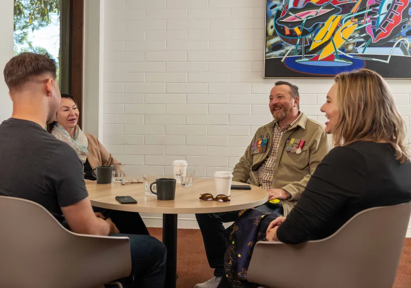 Four adults sitting around a table having a conversation over coffee in a casual indoor setting; one person wears military service medals.