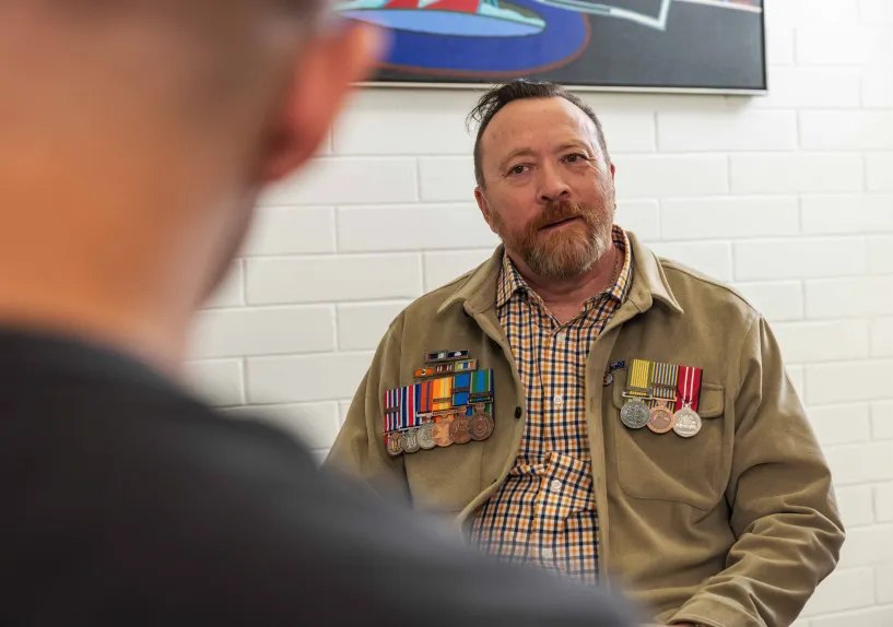 A person wearing multiple military service medals sits and listens during a conversation indoors.