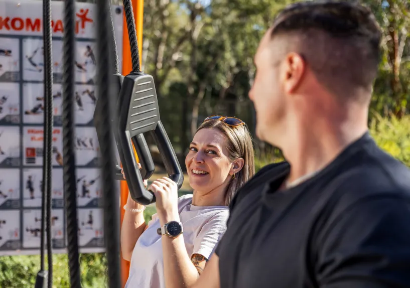 Two people using outdoor exercise equipment in a park setting.
