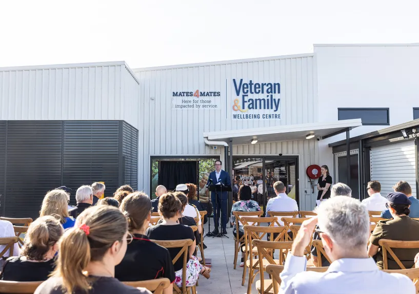Community gathering outdoors at the Veteran and Family Wellbeing Centre, with a speaker addressing an audience seated in front of the building.