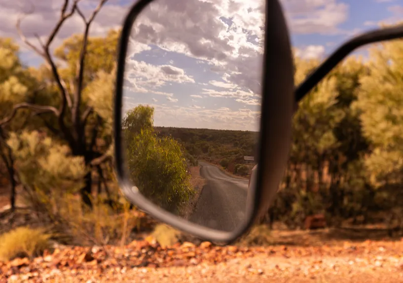 A winding road seen in a vehicle side mirror, with surrounding bushland and trees visible along the roadside under a partly cloudy sky.