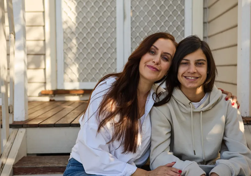 An adult and teenager sitting side by side on front steps, leaning close with an arm around each other.