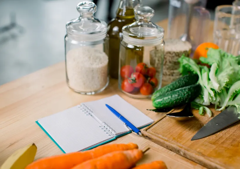 Fresh vegetables, pantry jars and a notebook with a pen arranged on a kitchen bench beside a chopping board and knife.