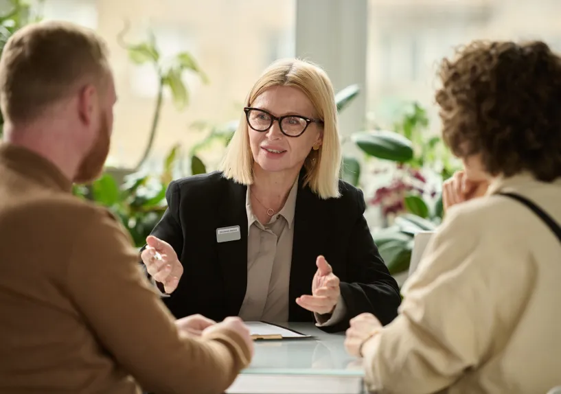 A person in a meeting explaining something to two others seated across a table, with documents in front of them.