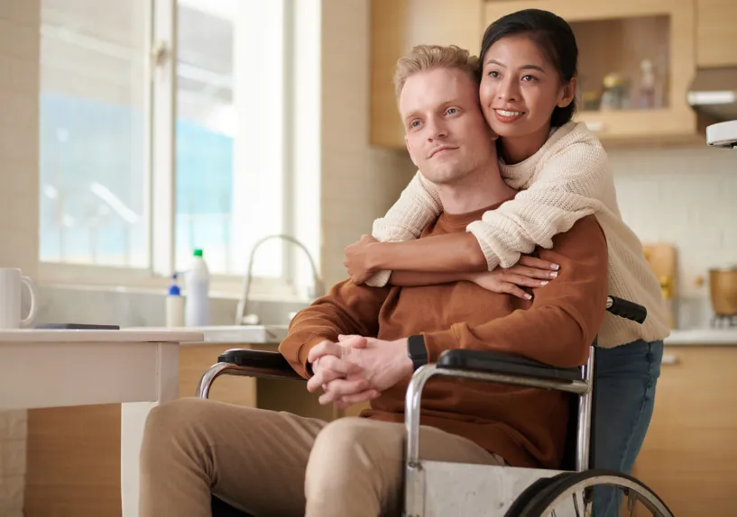 A person in a wheelchair sitting at a kitchen table while another person stands behind with arms around their shoulders.