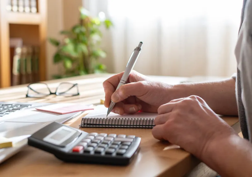 Close-up of a person writing in a notebook with a pen at a desk, with a calculator and laptop nearby.