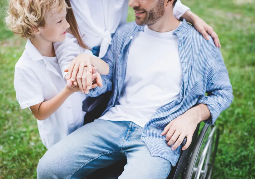 Close-up of a child and an adult in a wheelchair holding hands with another person, standing together outdoors.