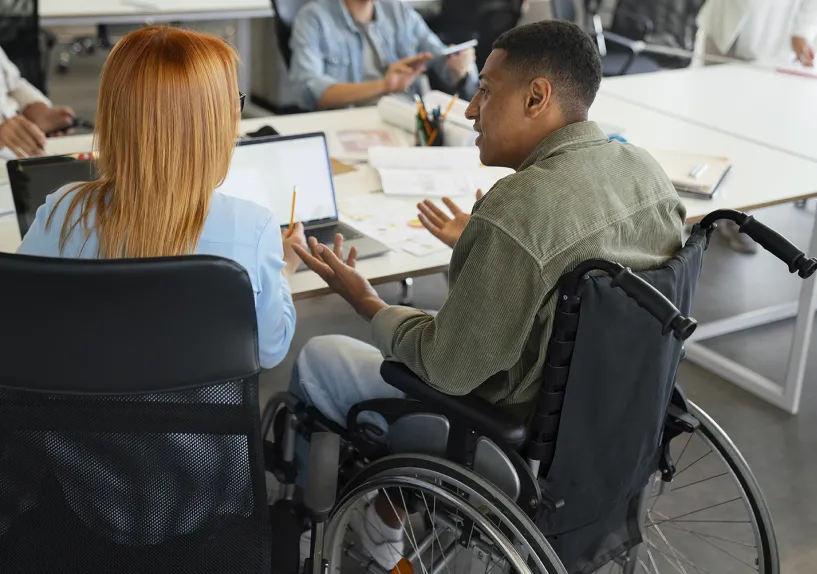 A person in a wheelchair sits at a desk in an office, engaged in conversation with other people around them.