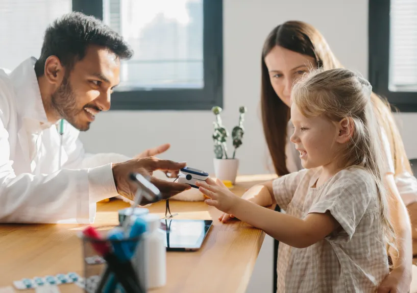 A doctor showing a medical device to a child at a table, with another adult sitting beside them.