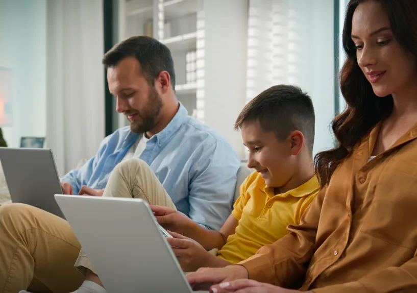 Two adults and a child sitting together on a couch using laptops and a tablet in a home setting.