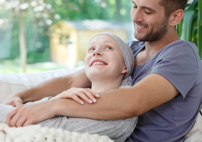 A person sitting on a couch, being hugged from behind by another person resting their arms around their shoulders.