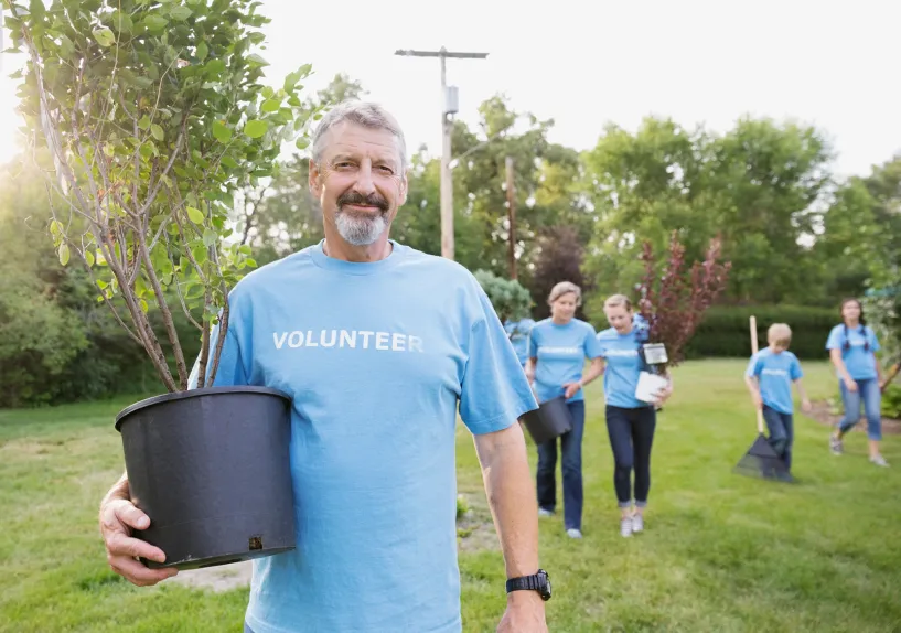 A person carrying a potted tree in a grassy outdoor area, with several volunteers in matching shirts walking behind with plants and tools.