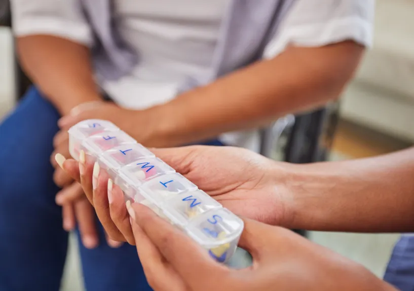 A person holding a labelled weekly medication organiser being passed to another person.