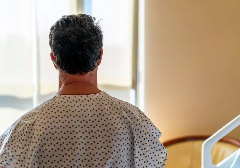 A person wearing a hospital gown sitting on a bed and looking out a window in a hospital room.