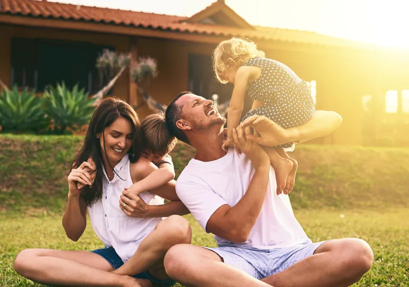 A family sitting on grass outdoors playing together, with one adult lifting a child while others sit nearby in front of a house.