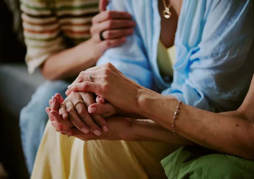 Close‑up of one person holding another person’s hands, with a third person resting a hand on their shoulder nearby.