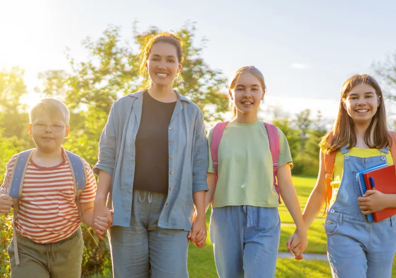 An adult walking with three children with backpacks, holding hands outdoors, with one child carrying a book.