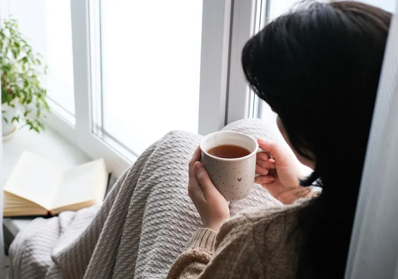 A person wrapped in a blanket sitting by a window, holding a cup of tea, with an open book nearby on the windowsill.