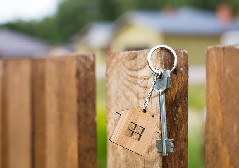 A key with a house-shaped keyring hanging on a wooden fence post.