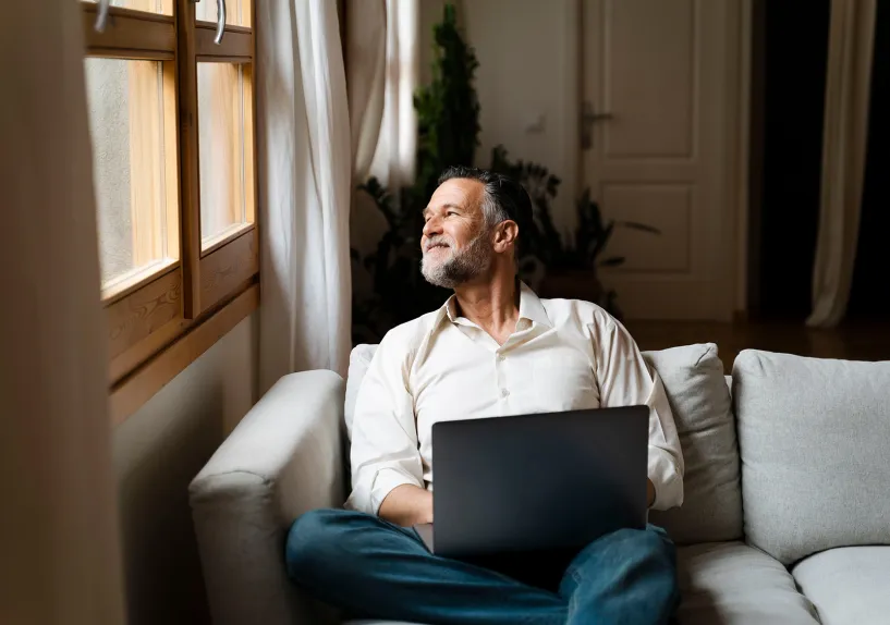 Man sitting on couch, looking out window with a laptop resting on his lap, 