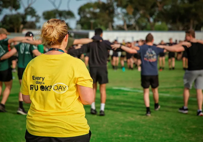 A woman wearing a yellow t-shirt which says Every day is R U OK? Day. A group of people are in the background linking arms.